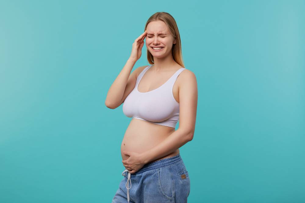 Upset Young Attractive White Headed Lady With Natural Makeup Keeping Her Eyes Closed While Having Strong Headache Being Pregnant While Posing Blue Background